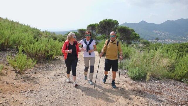 Group of senior friends hiking along trail in countryside, laughing and enjoying a wonderful hike together in a scenic natural landscape. Daytime, spring. Full-body, wide shot. 