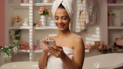 Hispanic beautiful woman in towel Arabian happy girl bathroom hotel smiling scrolling smartphone online shopping app cosmetics delivery home using mobile phone internet purchase beauty skincare bath