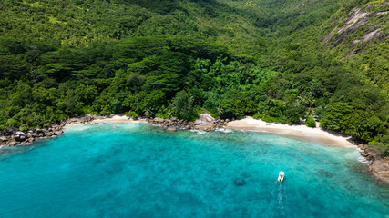 Granite rocks nestled in dense tropical forest next to a beach with clear turquoise water. Anse...