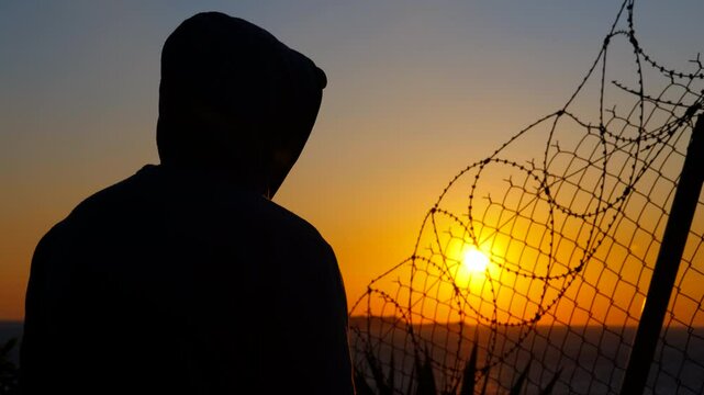 Man removing hood at sunset behind barbed wire. Hooded silhouette removing hood while standing before barbed wire fence, gazing at sunset sky with sense of freedom, hope, and new beginning emerging