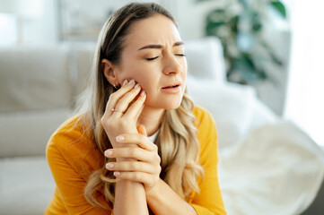 Upset caucasian woman in a yellow shirt sitting on a couch at home with eyes closed, holding her...