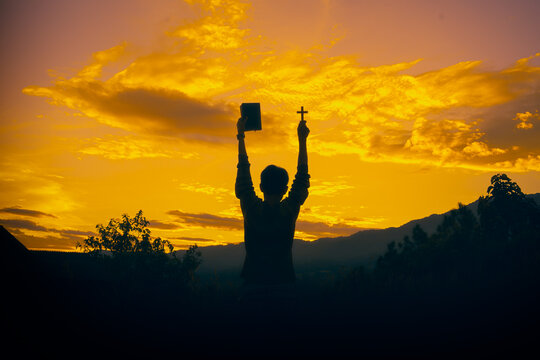 One human standing praying and holding christian cross whit bible for worshipping God at sunset background. christian silhouette concept.