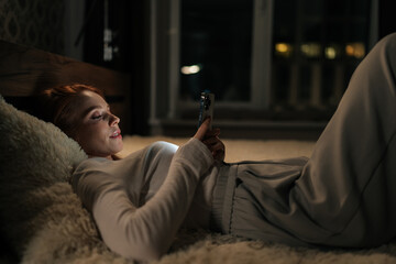 Side view of serene young woman lying in bed at night, scrolling browsing social media and messaging, face illuminated by soft glow of smartphone, creating cozy serene atmosphere, bokeh light. © dikushin