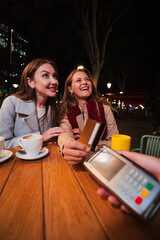 Vertical. Two Joyful Women Enjoying a Delightful Evening Out, Engaging in Cheerful Conversation While Preparing to Make a Payment with a Credit Card at a Cozy Cafe Table with Coffee Cups