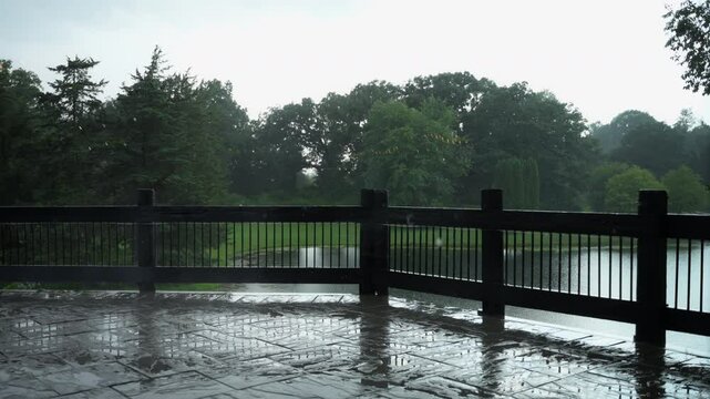 Rain falling on backyard with fancy wooden fence and green park background, on a wet summer partly sunny day. Big rain drops.