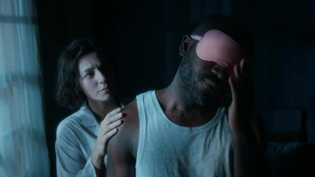 Young Caucasian woman looking worried, taking her sleepwalking African American husban by shoulder and waking him up in night