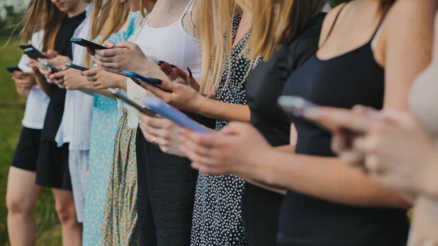 Group of young women are standing in a line outdoors, each looking down at their smartphones.