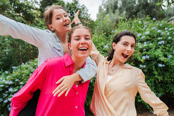 Three cheerful women smiling at the camera outdoors, enjoying a playful moment with one giving a...