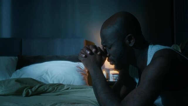 African American man kneeling by bed and praying with his hands clasped before going to sleep in his bedroom