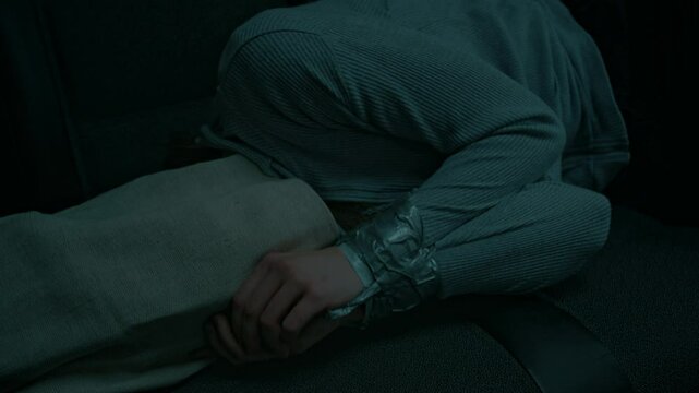 Lowkey medium shot of unrecognizable hostage lying in dark truck with sack on head and tied up hands, human trafficking and abduction