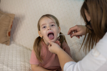 A female pediatrician examines the throat of a sick little girl with a wooden stick during a home...