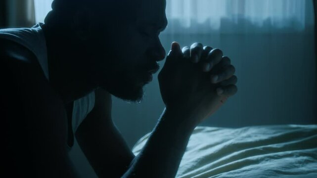 Close-up shot of African American man clasping his hands and saying prayers out loud with his eyes closed, kneeling by bed before going to sleep at night