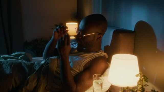 African American man taking off his glasses and putting away book before turning off bedside table lamp and falling asleep at night in hs bedroom