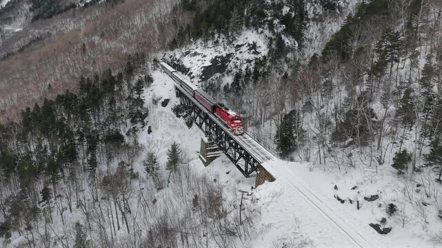 Train crossing trestle bridge in White Mountains of New Hampshire on snowy winter day. Aerial drone video filmed in Crawford Notch, NH.