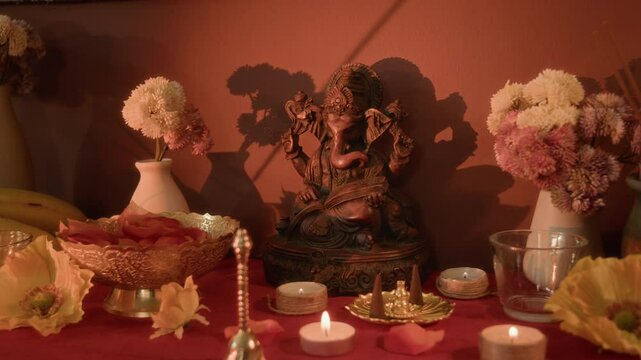 No people warm light shot of statue of Hindu god Ganesha on home altar surrounded by flowers, candles, bell and traditional decor on red tablecloth, changing focus