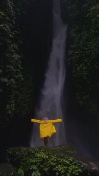 Vertical: Blonde girl at waterfall flowing in Bali raises hands up. Hiking, Woman in a yellow raincoat or jacket in the tropical jungle enjoys a vacation or trip, lifts palms. Rainy season, Indonesia.