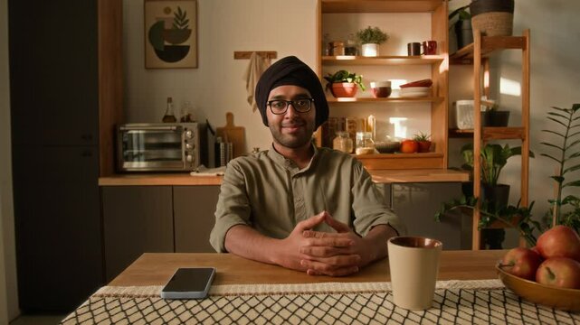 Medium warm light shot of smiling young adult Indian man in glasses, traditional black turban and khaki kurta sitting at dining table in modern domestic kitchen and looking at camera