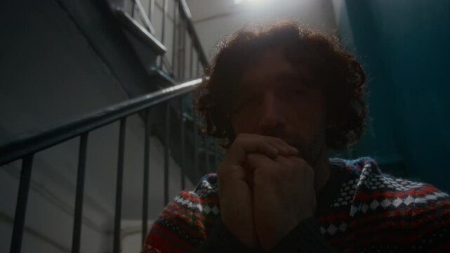 Closeup of apathetic or anxious man with curly hair and stubble, wearing sweater, sitting alone in stairway landing and looking around indifferently,