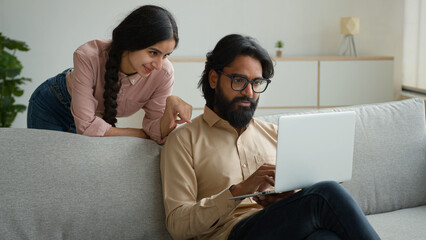 Arabian Indian couple on couch sofa use computer laptop shopping online internet store happy multiracial wife husband smiling lovely customers woman and man search goods purchase on website at home