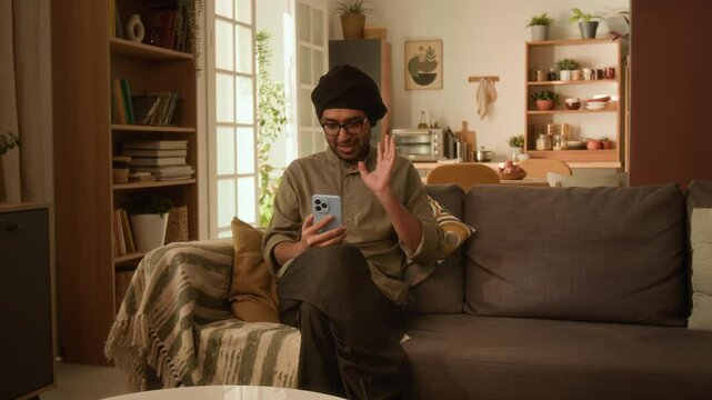 Full shot of pleasant Indian man in glasses, traditional kurta and turban sitting on sofa at home and video calling friends on mobile phone, warm light