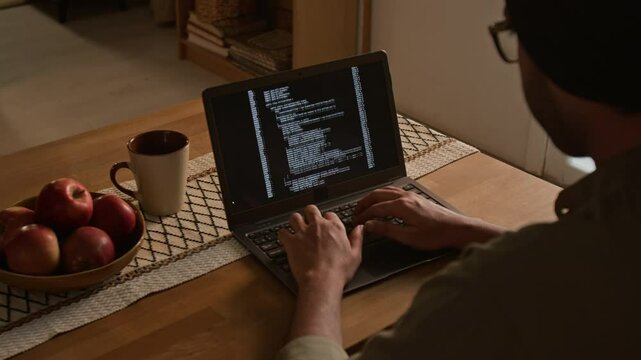 Over shoulder shot of anonymous man with covered head typing machine code on wireless laptop sitting at dining table and working remotely from home