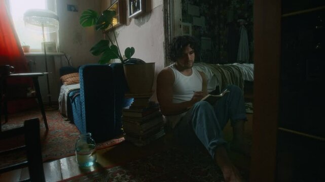 Full shot of apathetic Caucasian gen Y man in undershirt and jeans sitting on floor in worn out old apartment and writing thoughts in diary, spending meaningless days alone