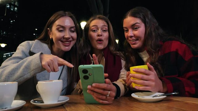 Group of young women smiling sharing media with an cellphone. Three girls looking to the smartphone on a coffee shop, restaurant or bar terrace. Female buying on a market place using a mobile phone