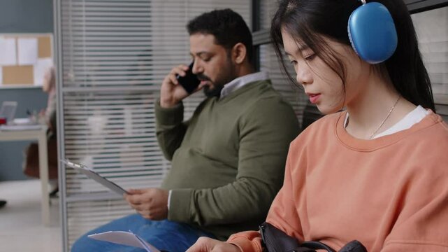 Side chest up shot of young Chinese girl in headset preparing for visa interview sitting in waiting room with man in embassy