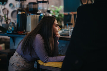 A young woman is seated in a cozy cafe, leaning on the counter, and gazing thoughtfully. The ambiance is warm and inviting, with soft lighting and visible coffee-making equipment.