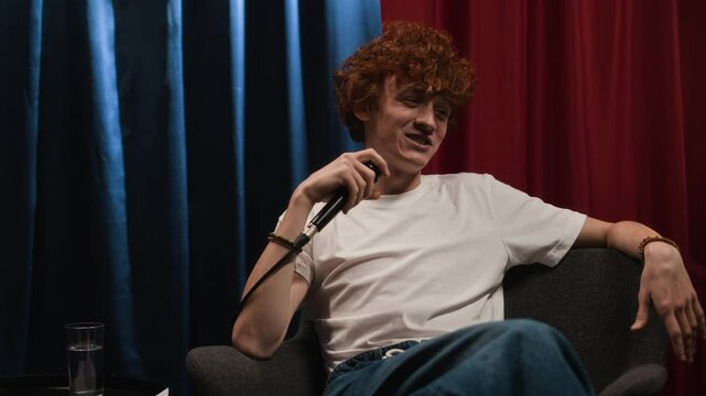 Full medium low key shot of confident and cheerful male Caucasian stand-up comic with ginger hair, wearing white t-shirt and jeans, sitting casually in armchair and telling jokes into microphone