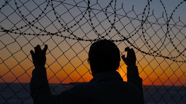 Prisoner looking at sunset through barbed wire fence. Silhouetted prisoner gazing through barbed wire fence, while watching radiant sunset symbolizing potential freedom