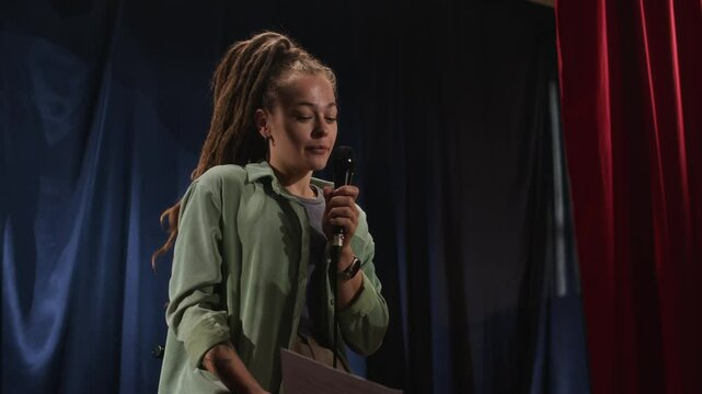 Low angle shot of Caucasian female standup comedian with dreadlocks dressed in dark green shirt and lilac t-shirt, performing on stage facing audience, slightly nervous and peeking at her text