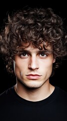 Close-up portrait of a young man with curly brown hair against a black background. Intense gaze and serious expression.
