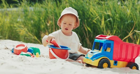 The boy is playing on the beach next to the water, pouring sand into a bucket using a shovel, next to him the rest of the toys, the child is focused on the activity