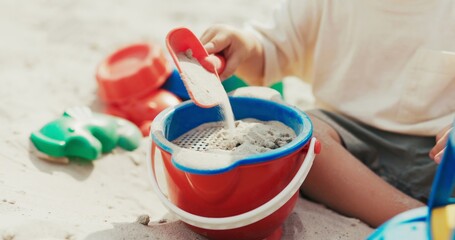 A child plays in the sandbox, a little boy spends his childhood in the backyard, pouring sand from a shovel through a strainer into a bucket, playing by himself, building castles