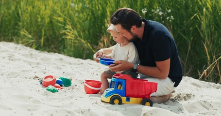 Naklejka premium A dad is playing with son in a sandbox, on the beach, toys are lying around them, a man is holding a small child on his lap, showing him how to pour dry sand from a strainer into a bucket
