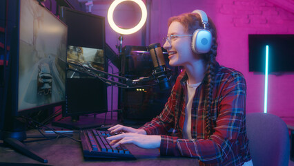 Happy gen z influencer blogger streamer teen woman girl female gamer player headphones neon light play computer video game racing cyber sport competition online stream gaming addict playing keyboard © Yuliia