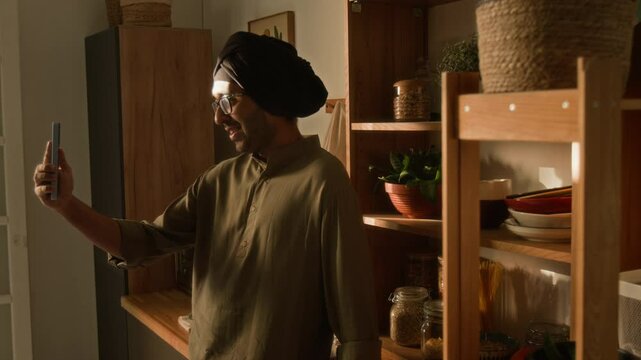 Side warm light shot of young adult Indian man in glasses, traditional turban and khaki kurta talking during mobile video call at home, connecting with family or friend