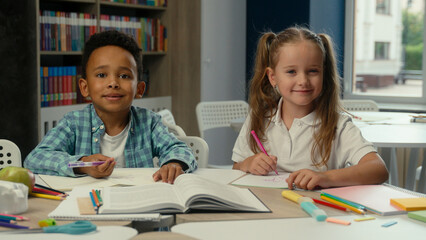 Fototapeta premium Cute little children multiethnic kids African boy Caucasian girl school pupils students classmates friends schoolboy schoolgirl in class together studying learning education smiling write draw at desk