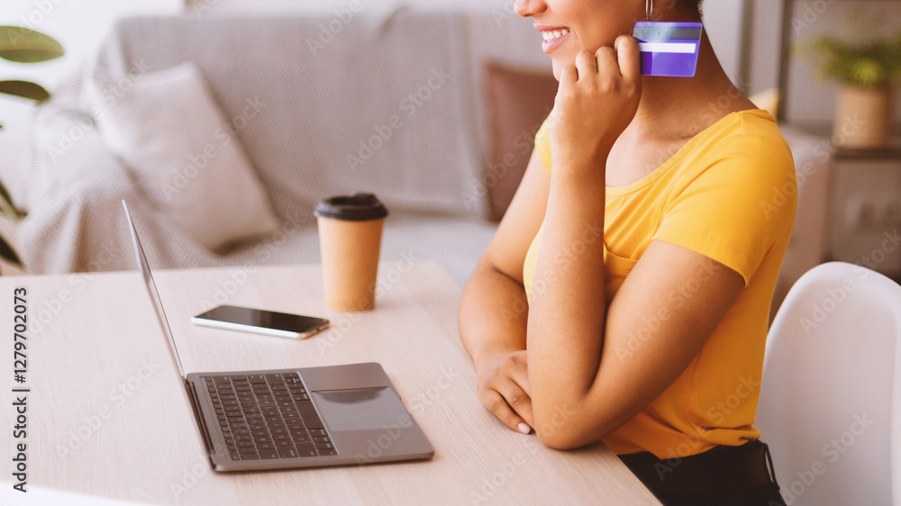 Poster Online Store And Internet Banking Concept. Side view portrait of unrecognizable smiling black lady holding credit card in hand and using laptop, sitting at table with phone and coffee, looking at pc
