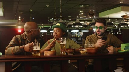 Medium lowkey shot of three diverse friends in green hats sitting together at bat counter and drinking beer, hanging out in evening on St Patricks day