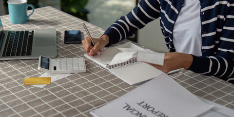 Individual taking notes for financial planning and bills while using a calculator and reviewing...