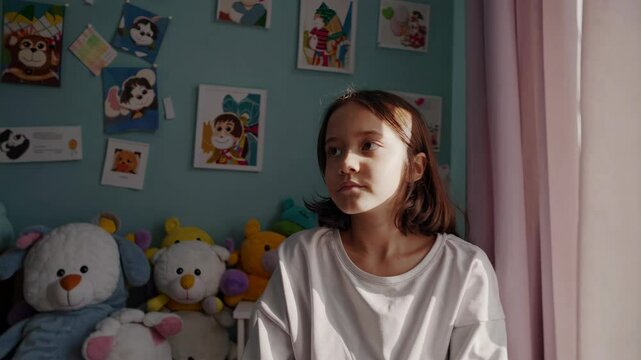 A young girl sits in a colorful room with toys and pictures, captured in a video from a low angle, highlighting a thoughtful expression.
