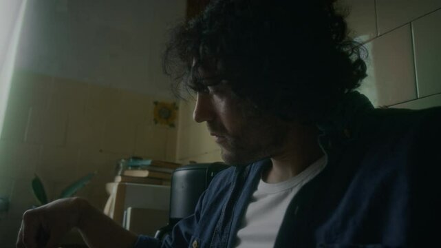 Side tilt shot of bearded Caucasian man sitting at kitchen table and leaving his breakfast untouched, indifferently looking out of window