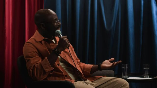 Full medium low key shot of confident African American male stand-up comedian in glasses and orange shirt sitting in armchair with mic on stage and telling jokes during night performance in club