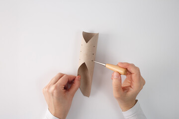 DIY craft process with hands using a pointed metal tool to punch holes in a cardboard tube. Paper roll shaped with petal-like ends for creative handmade project, possibly for decoration or assembly