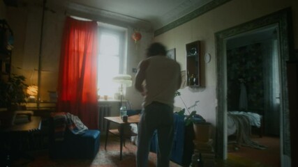 Full shot of restless and depressed man in undershirt and jeans wandering around room in shabby flat, reading letters and throwing them away, feeling alone and unwanted