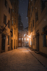 A snowy cobblestone street in Riga's old town, flanked by historic buildings with warm lighting. A tall church spire is visible in the background.