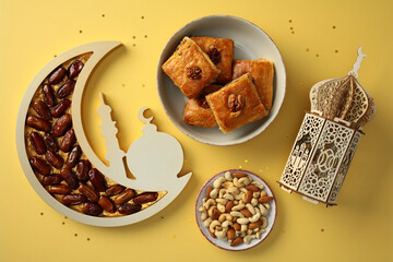 Ramadan celebration concept. Dates in a crescent moon tray, baklava on a plate, mixed nuts, and a decorative lantern on a yellow background with star confetti.