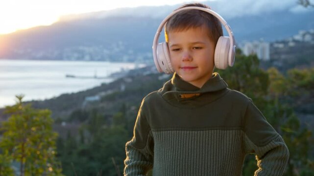 A boy stands in the mountains, against the backdrop of the city and the setting sun on a spring evening. He listens to music through headphones.